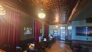 Een restaurant met houten vloeren, rode gordijnen en een kroonluchter. Er staan tafels en stoelen, en een bar met comfortabele zitplaatsen.