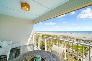 Un balcon avec vue sur la plage, des meubles blancs et un fauteuil en osier.