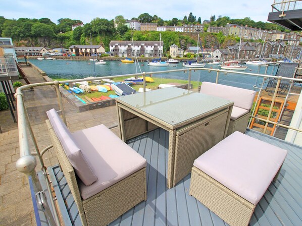 A deck with wicker furniture and a table, overlooking a marina with boats and houses in the background. - Dros Y Dwr (Porthmadog)
