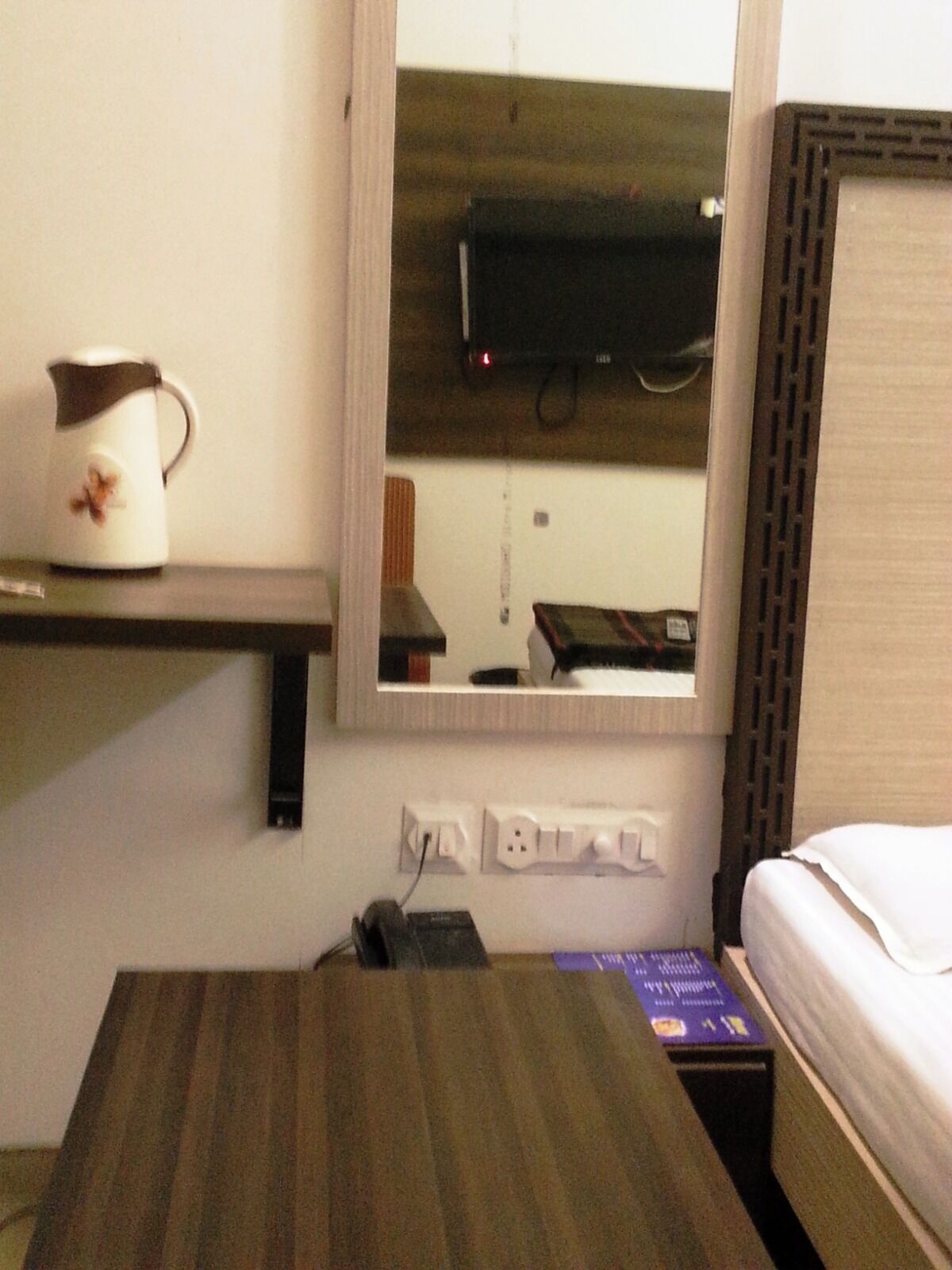 Une chambre d’hôtel avec un lit, une petite table, une bouilloire et un miroir dans lequel se reflètent un téléviseur et une fenêtre.