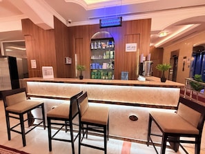A hotel reception area with a wooden reception desk, a digital screen, and a 'Bonjour Cafe' sign. There are four chairs in front of the desk and a potted plant on the counter. - Alfursan Almotahida Serviced Apartments Alsalama Dist (Jeddah)