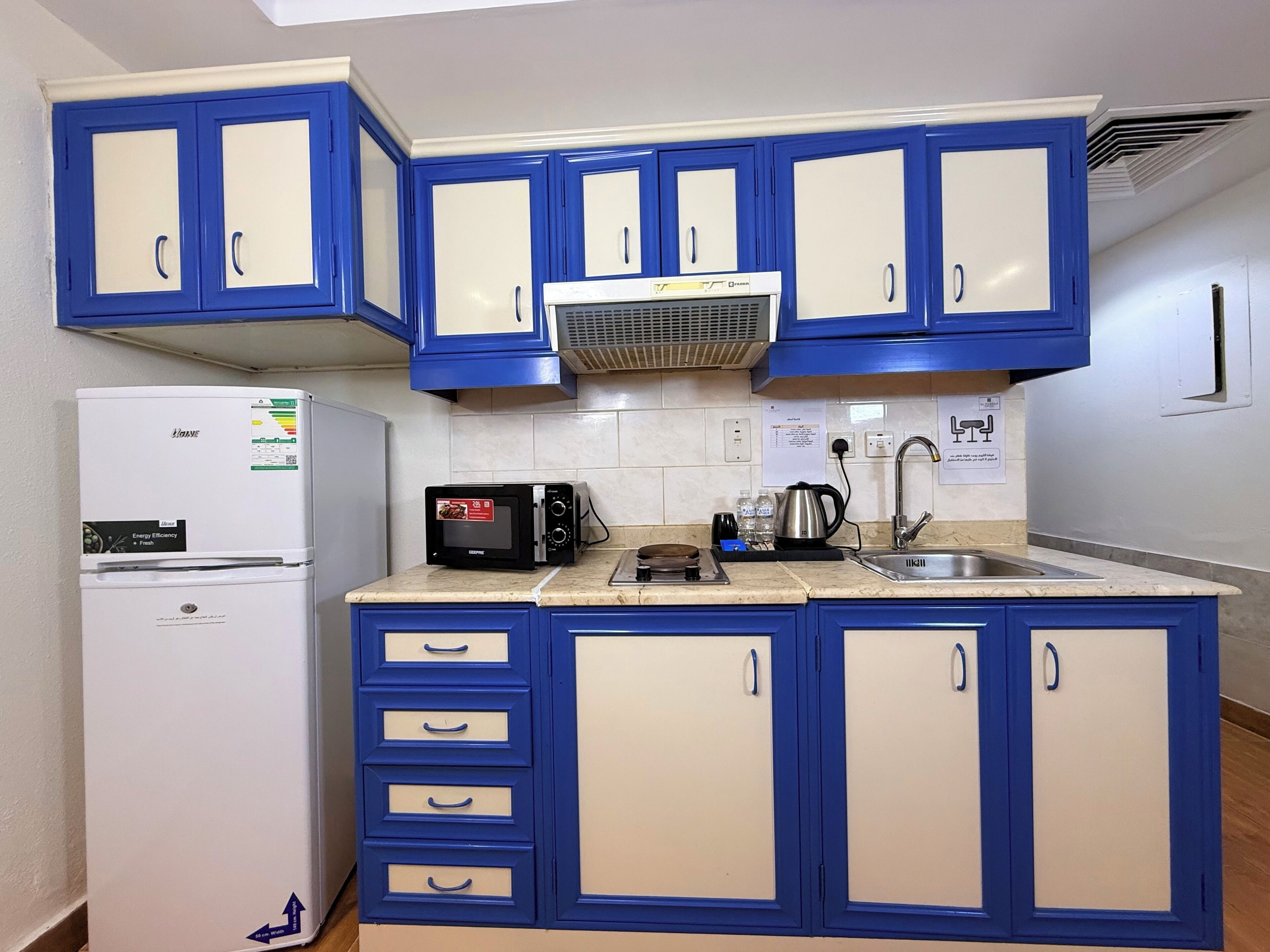 A compact kitchen with blue cabinets, a white refrigerator, a microwave, a kettle, and a sink.