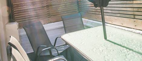 Une terrasse avec une table et des chaises en verre, un parasol noir et un mur de briques en arrière-plan.
