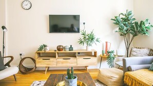 Una sala de estar con un mueble de madera para el entretenimiento, un televisor de pantalla plana, un sillón de mimbre, un sofá y varias plantas.