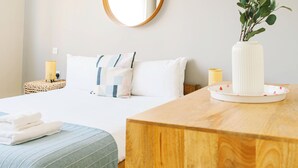 Habitación de hotel con una cama bien tendida, un espejo redondo en la pared, una mesita de noche con lámpara y un escritorio de madera con un jarrón de plantas verdes.