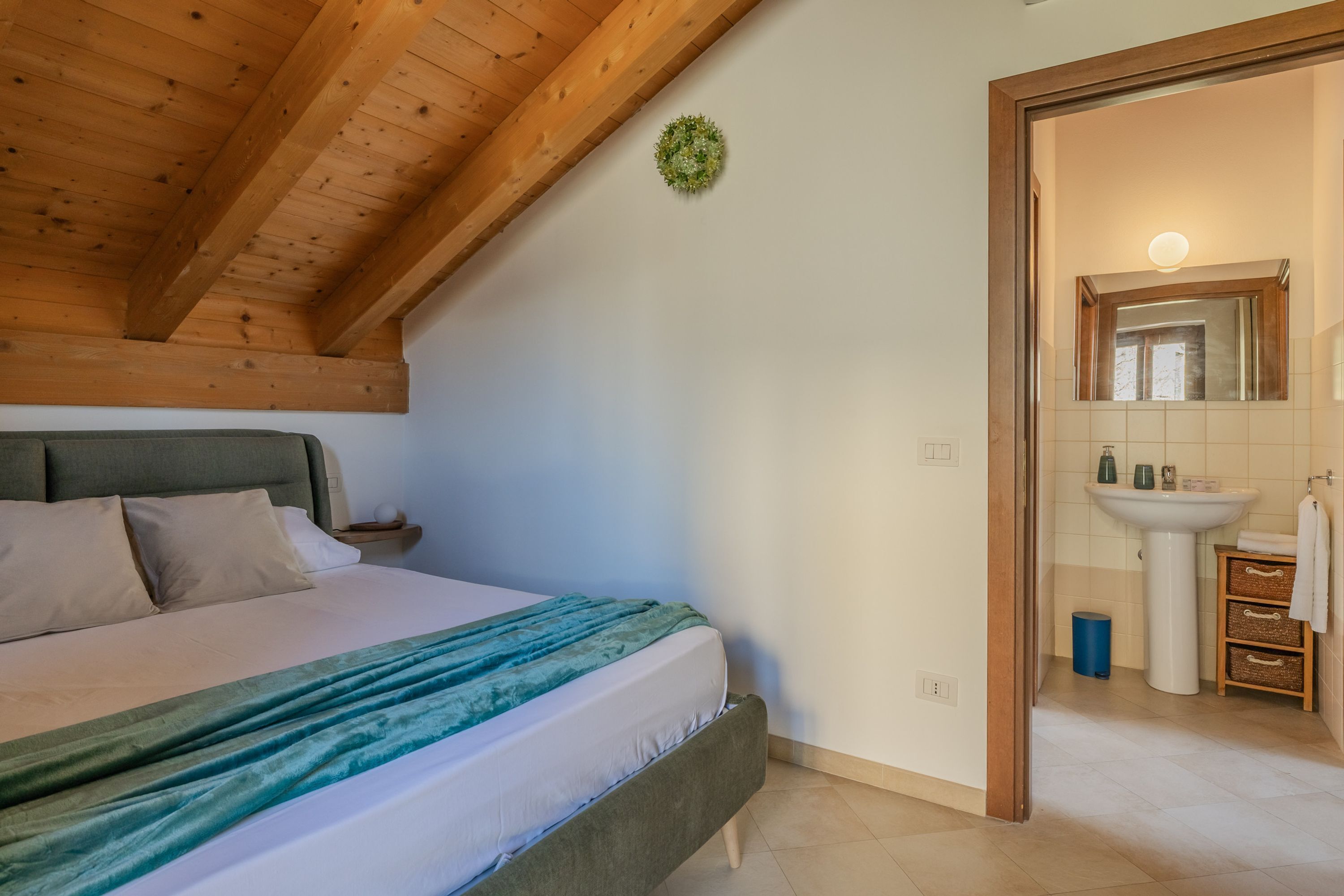 A bedroom with a bed, a nightstand, a wall-mounted light, and a bathroom with a sink and mirror.
