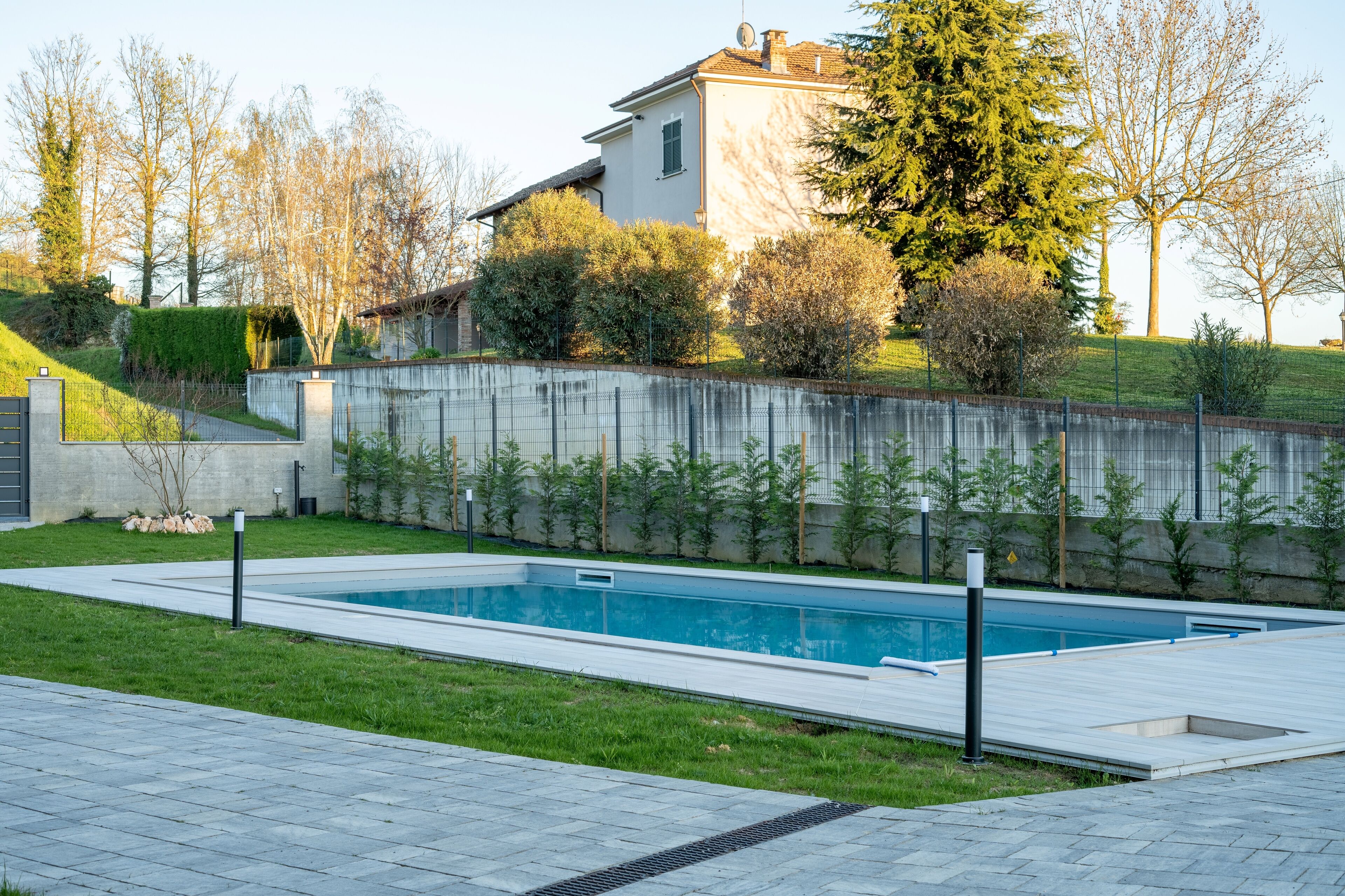 A rectangular swimming pool with a surrounding paved deck, a low stone wall, and a grassy area with trees and shrubs.