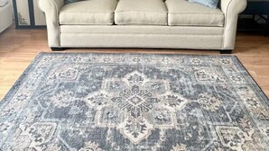 Uma sala de estar com um sofá bege, duas almofadas azuis, um tapete estampado, um quadro emoldurado retratando uma paisagem costeira e uma pequena prateleira com objetos decorativos.