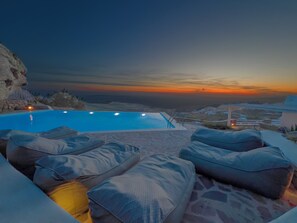 Una piscina infinita con vistas al atardecer, asientos exteriores con cojines y un patio de piedra.