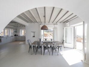 Une cuisine moderne avec des meubles blancs, un coin repas doté d’une grande table et de chaises, et une vue sur l’extérieur grâce à une porte vitrée.