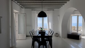 A dining area with a table and chairs, a hanging light fixture, and a view of the sea through large windows. - Villa Prive 88 Mykonos (Mykonos)