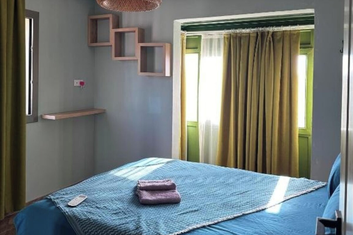 A bedroom with a bed, a hanging light fixture, a window with curtains, and a wall-mounted shelf.