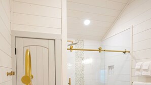 Un baño con cabina de ducha de vidrio, un banco de madera, un perchero blanco y suelo de madera.