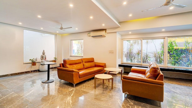 A modern living room with a large sofa, a smaller sofa, and two round tables. There is a ceiling fan, recessed lighting, and a large window with a view of greenery outside.