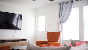 A living room with a flat-screen TV, a sofa, an orange armchair, and a window with curtains.
