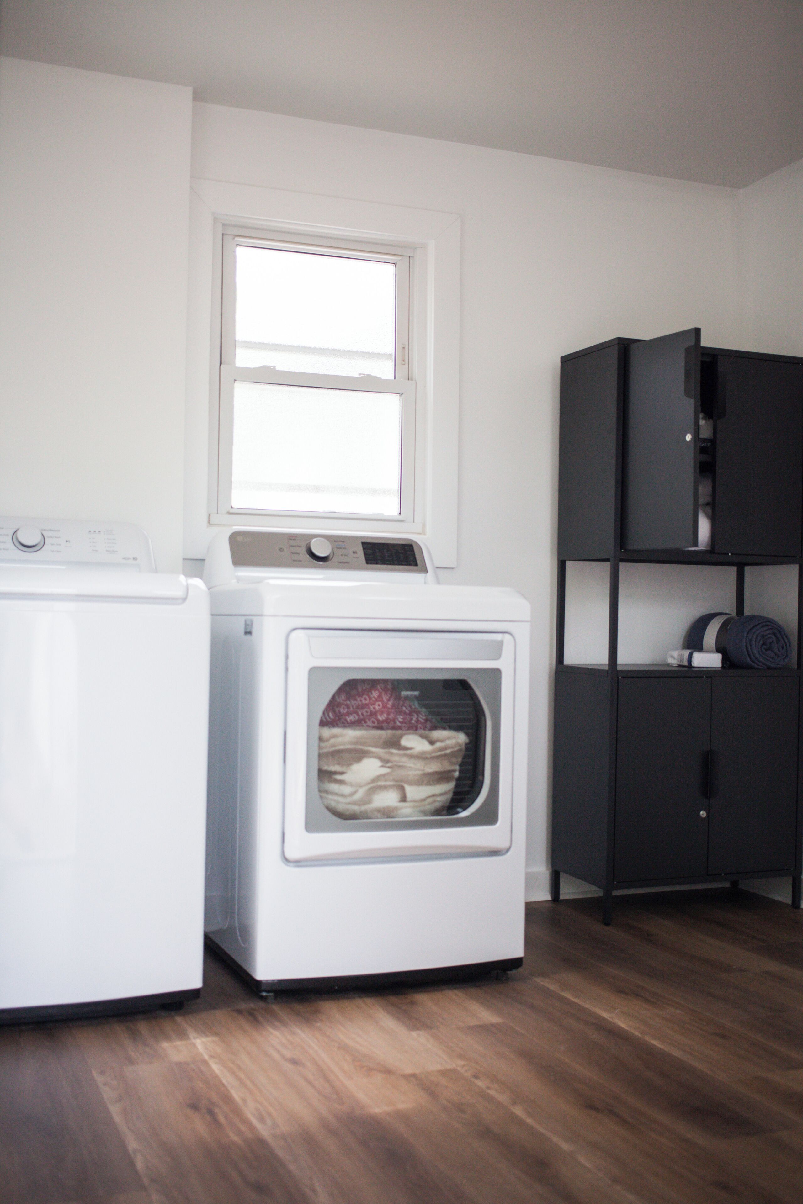 A laundry room with a front-loading washer and dryer, a window above the dryer, and a dark-colored shelving unit to the right.