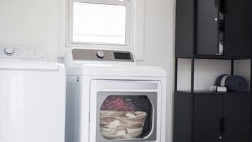 A laundry room with a front-loading washer and dryer, a window above the dryer, and a dark-colored shelving unit to the right.