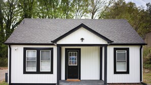 A small, single-story house with a black front door, two windows on the left, and a small porch.