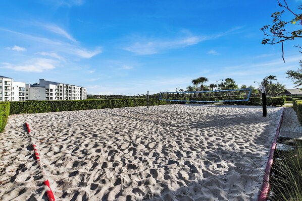 A sandy beach area with a volleyball net, surrounded by buildings and greenery.