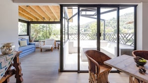 Un intérieur moderne avec un plafond en bois, un canapé bleu, des chaises blanches, une table à manger sur laquelle se trouve un saladier de fruits, et une vue sur un balcon avec une balustrade.