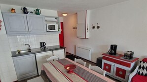 Uma cozinha compacta com armários brancos, micro-ondas e máquina de café. Há uma mesa de jantar com um centro de mesa vermelho e um banco.