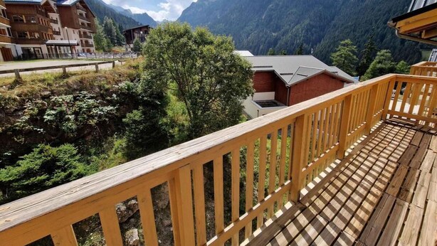 Un balcón de madera con vistas a un paisaje montañoso, un edificio de techo inclinado y un cielo despejado.