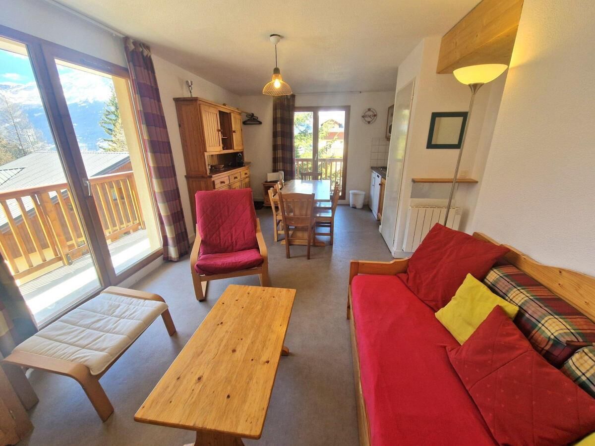 A cozy living room with a red sofa, a wooden table, and a red armchair. There is a balcony with a wooden railing and a view of the mountains.