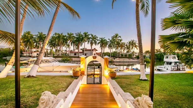 A wooden pathway leading to a gated entrance with a decorative archway, surrounded by palm trees and a body of water.