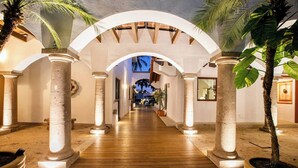 A corridor with white arches, columns, and wooden flooring, leading to a pool area with palm trees.
