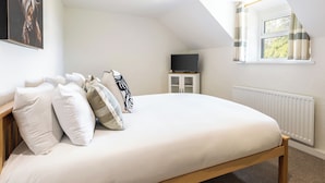Quarto com teto inclinado, cama com roupa de cama branca, televisão em um suporte e janela com cortinas.