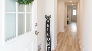 Un pasillo con piso de madera, una puerta blanca adornada con un arreglo verde, y un cartel que dice 'WELCOME'.