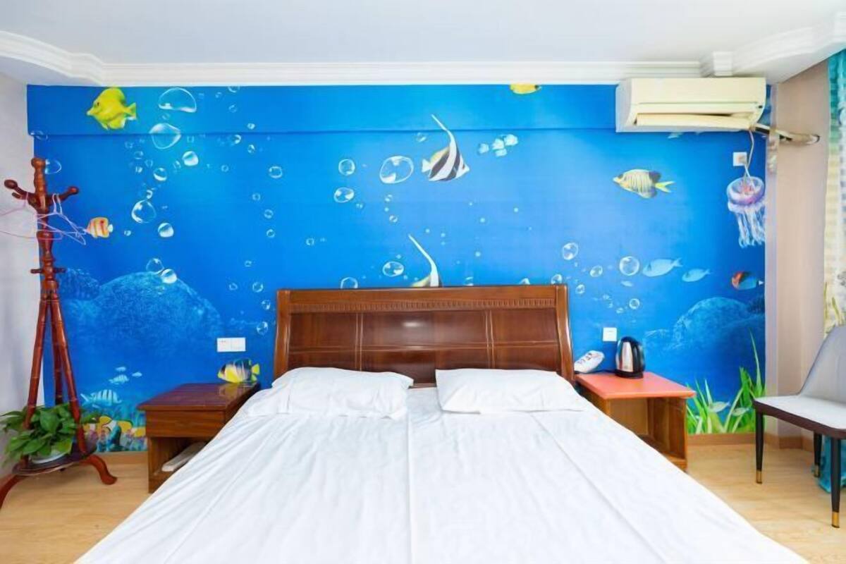 A bedroom with a blue ocean-themed mural, a wooden bed, a bedside table, a chair, and a plant.