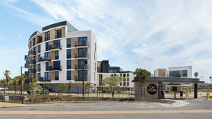 A modern multi-story building with balconies, a parking area, and a clear sky.