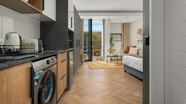 A modern kitchen with a built-in washer and dryer, a dining area with a table and chairs, and a bedroom with a bed and a window.
