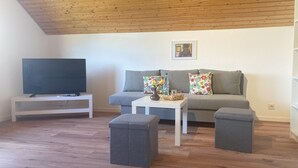 Un salón con techo de madera, un sofá gris con almohadones de colores, una mesa de centro blanca y un televisor de pantalla plana sobre un mueble blanco.