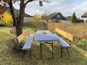 Un área de estar al aire libre con una mesa a cuadros y bancos, rodeada por una valla y árboles.