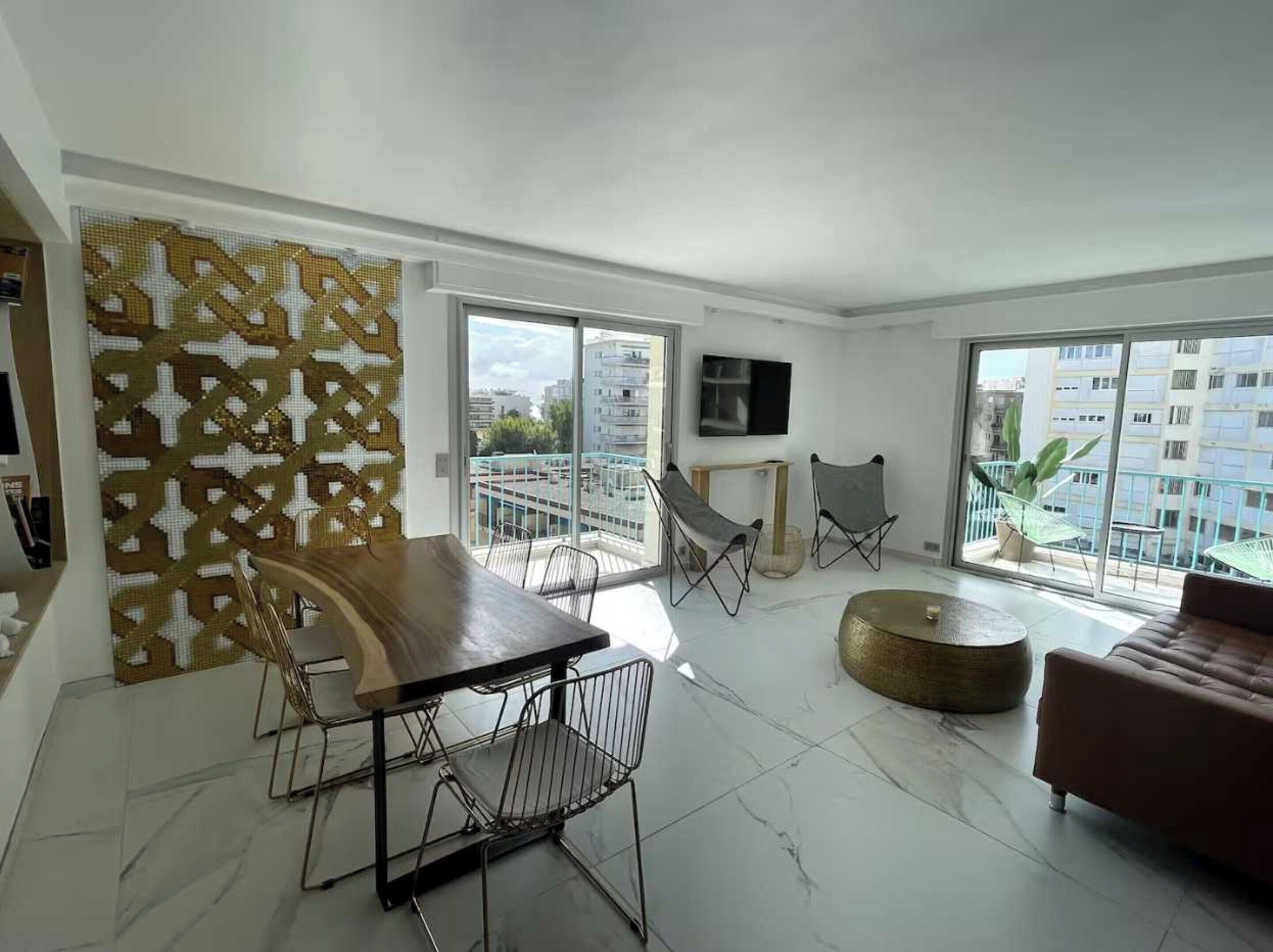 A modern living room with a dining area, large windows, and a balcony with a view of buildings.
