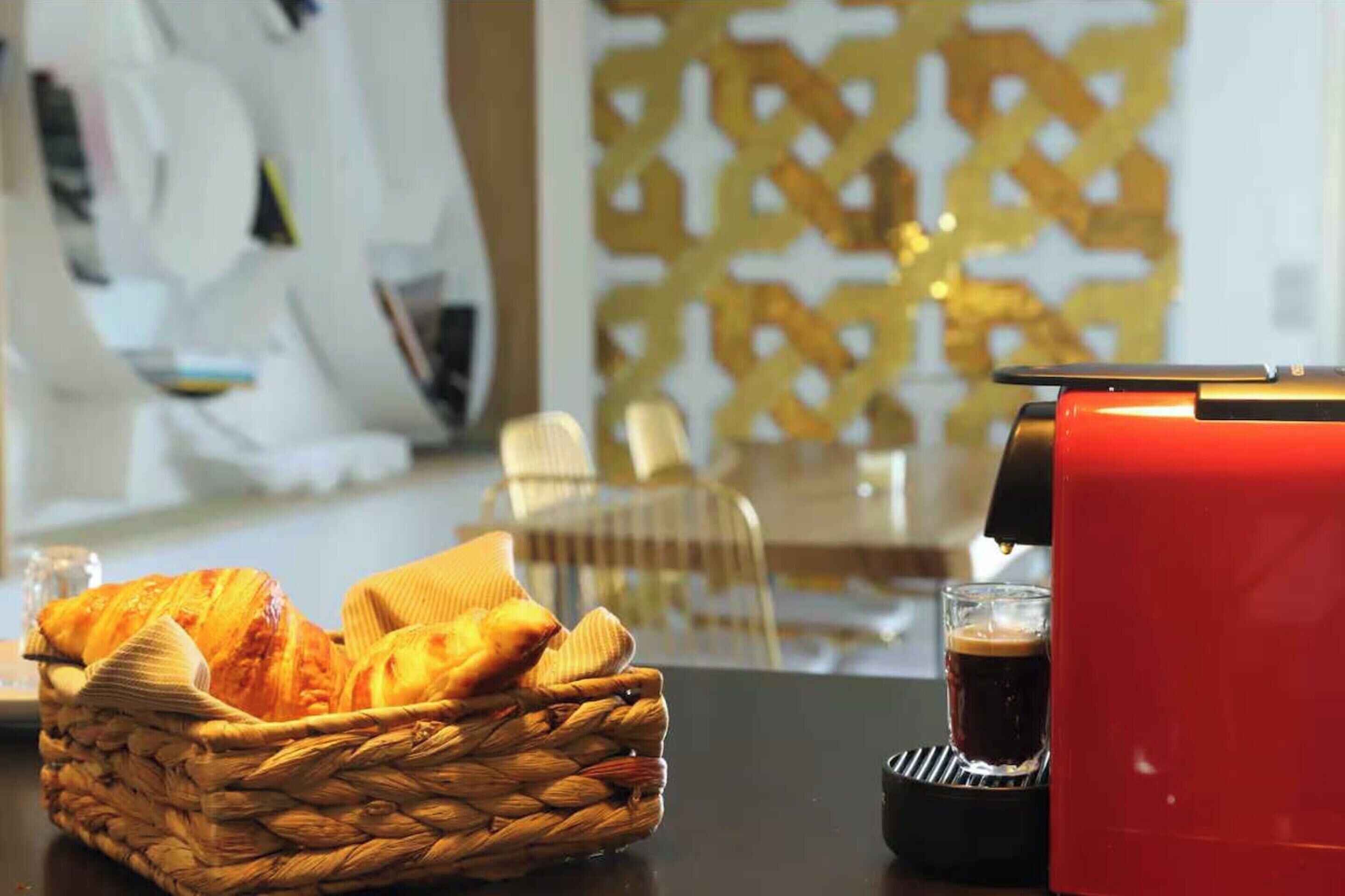 Ein Korb mit Croissants auf einem Tisch, eine rote Kaffeemaschine und eine Tasse Kaffee.