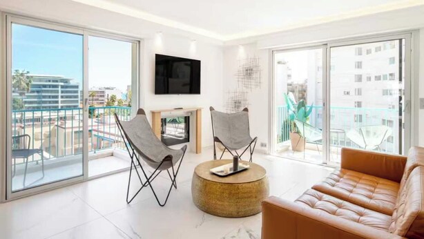 Een moderne woonkamer met een groot raam, een flatscreen televisie, twee stoelen, een rond poefje en een leren bank.