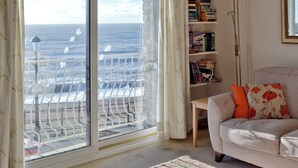 A living room with a large window overlooking the sea, a bookshelf, and a sofa with decorative pillows.