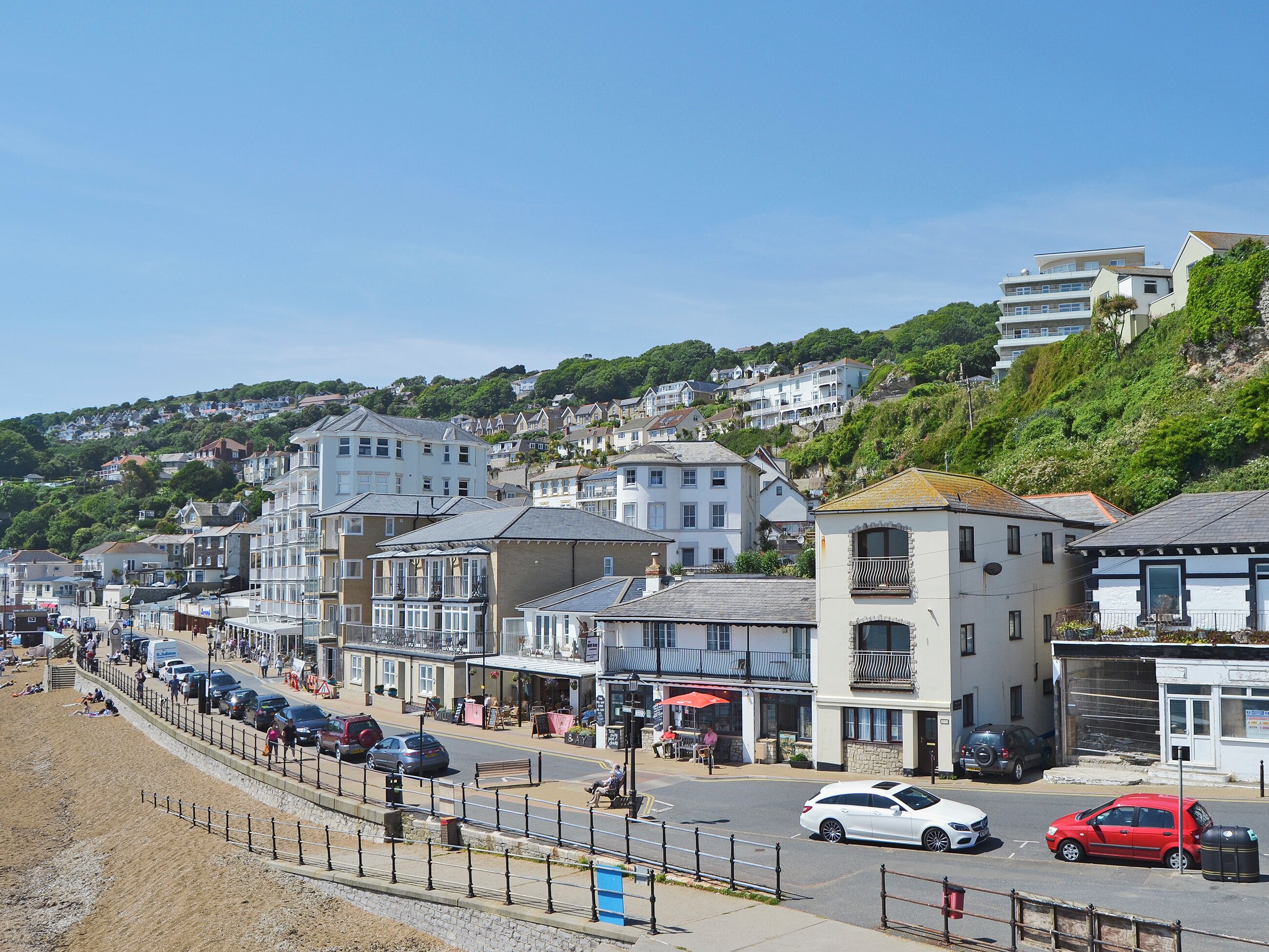 A coastal street lined with multi-story buildings, some with balconies, and a sandy beach area with a fence.