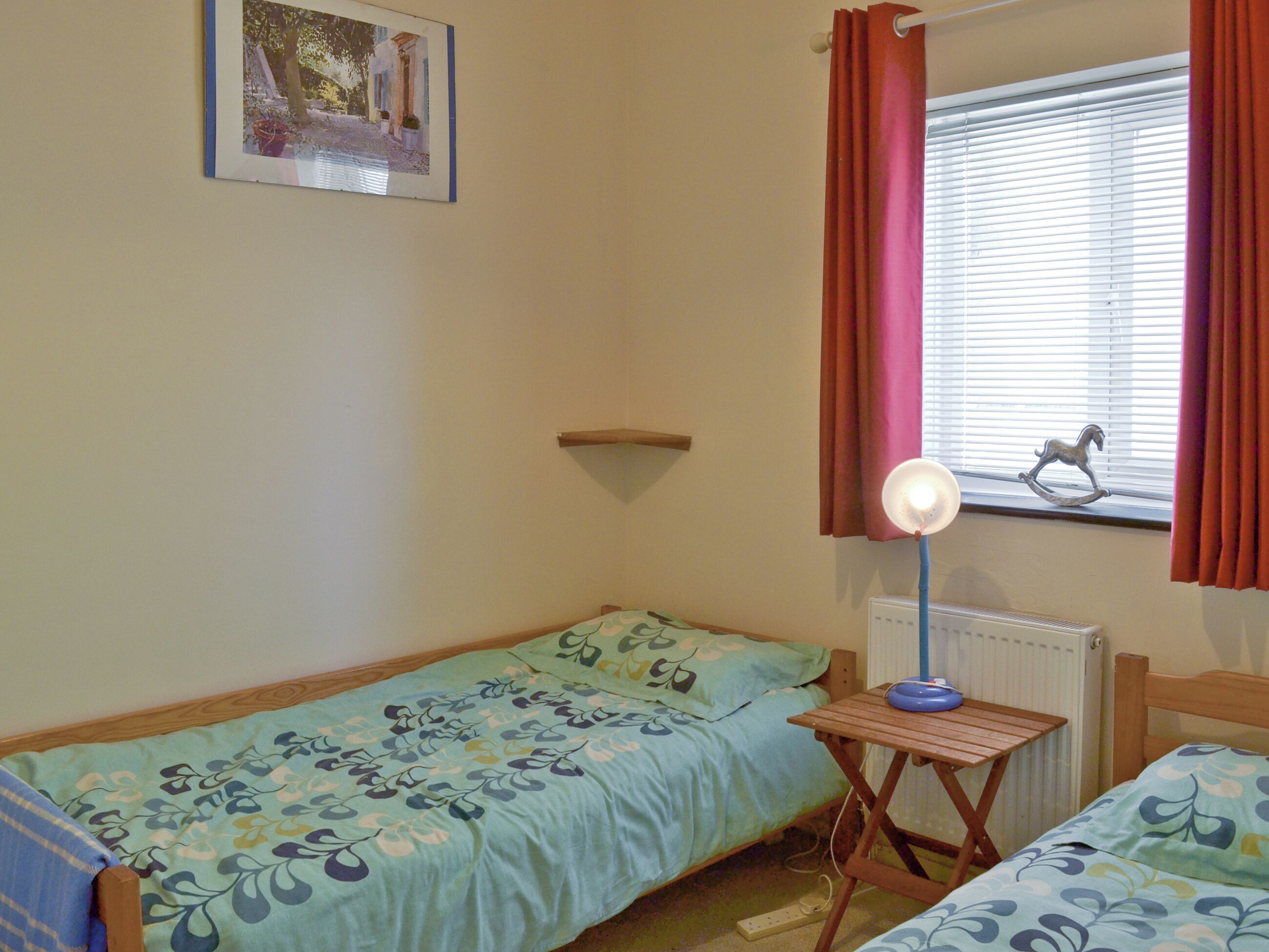 A small bedroom with two single beds, a bedside table with a lamp, a radiator, and a window with curtains.