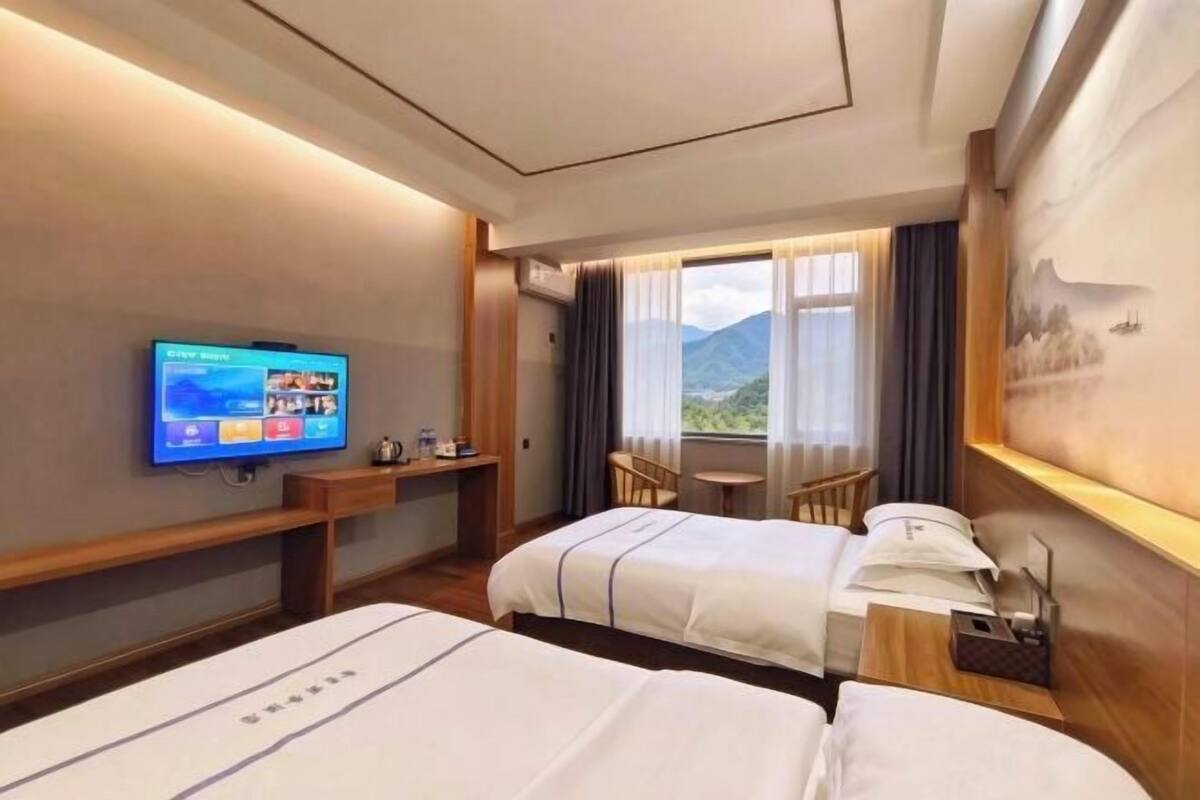 A hotel room with two beds, a TV, a desk, a chair, and a window with a mountain view.