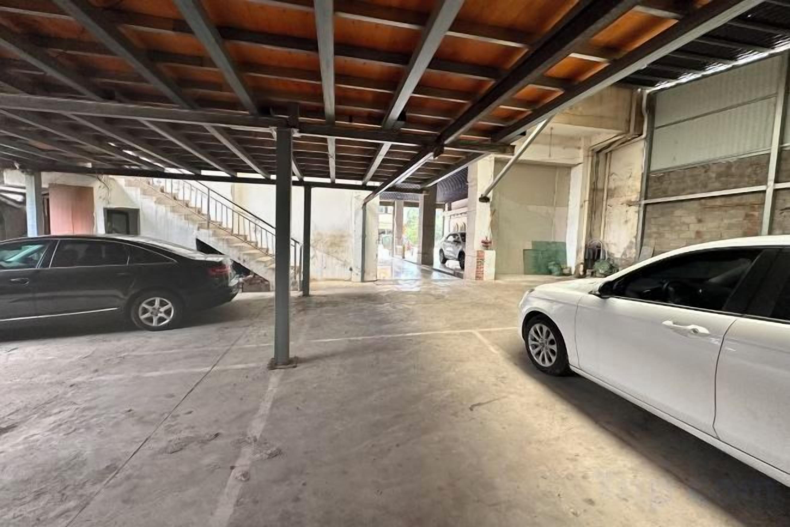 Ein überdachter Parkbereich mit zwei Autos, eine Treppe zu einer oberen Ebene und ein Gebäude mit einem großen Fenster.