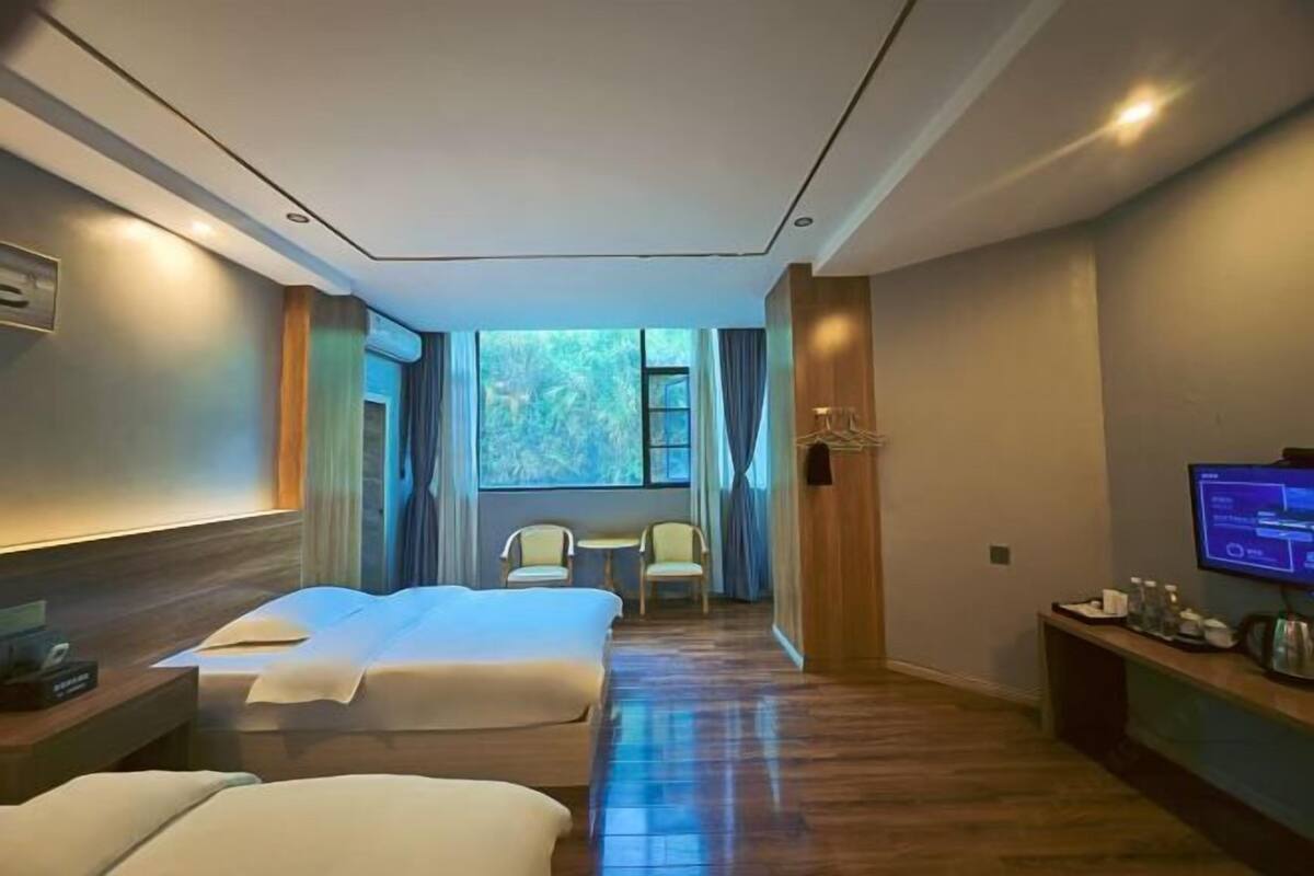 A hotel room with two beds, a TV, a desk with a computer, and a view of greenery outside.