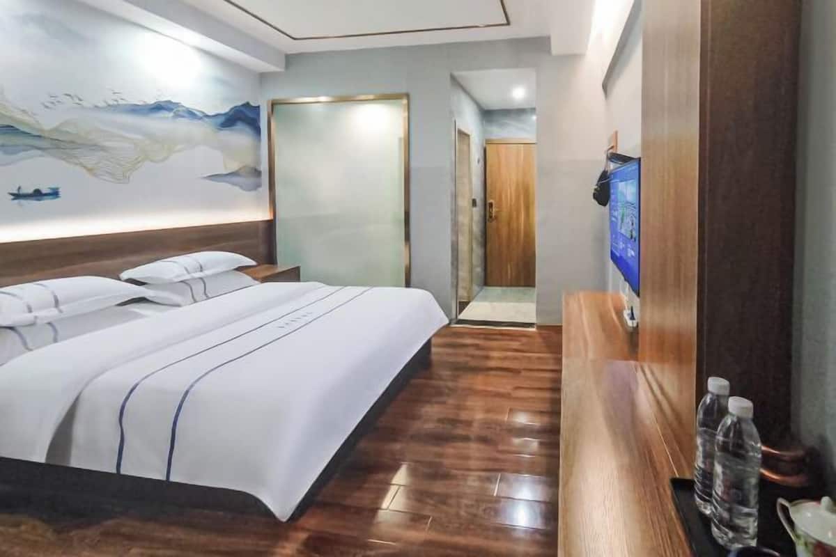 A hotel room with a large bed, wooden flooring, a painting on the wall, and a television on the right side.