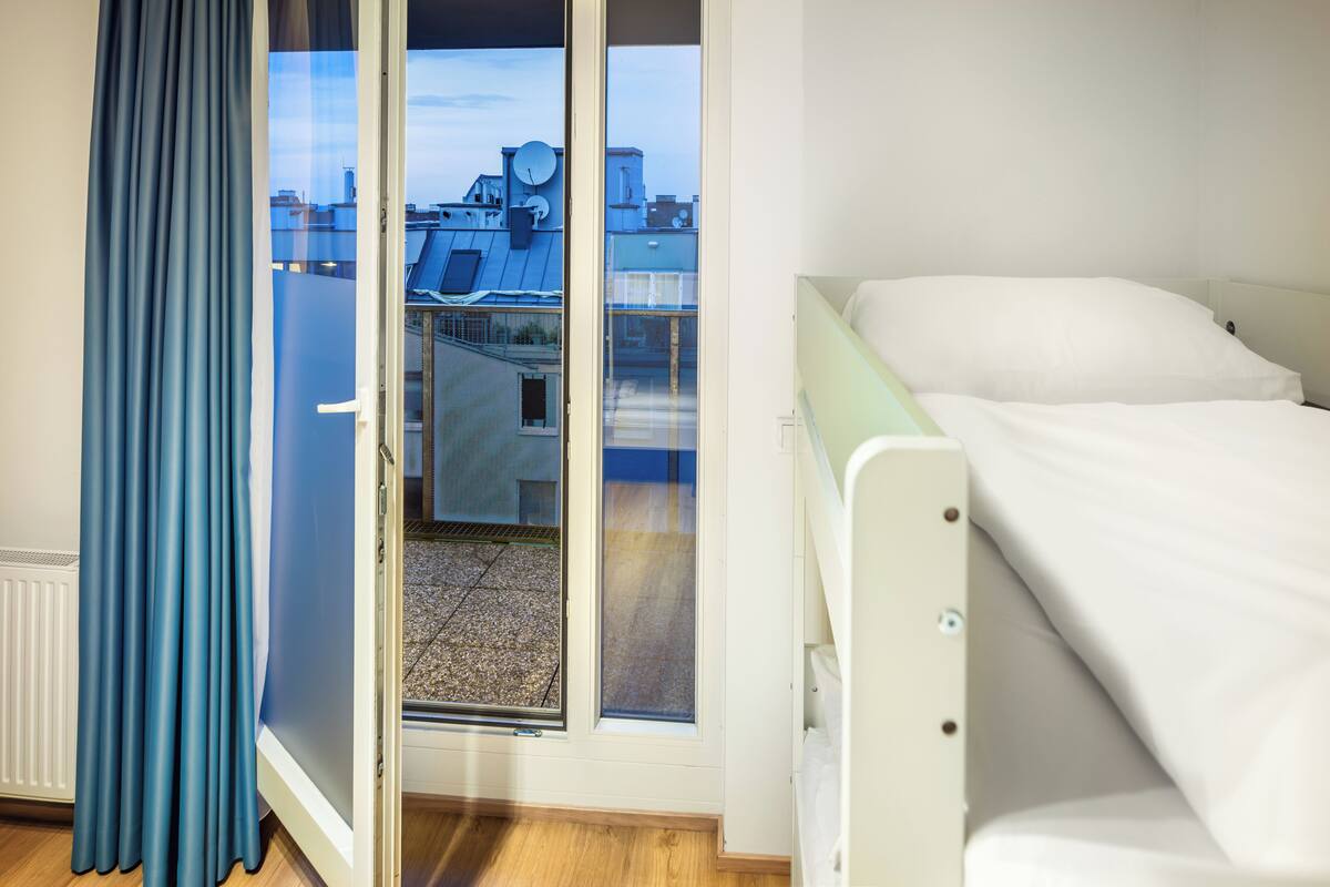 A room with a bed, a window with a view of buildings, and a curtain.