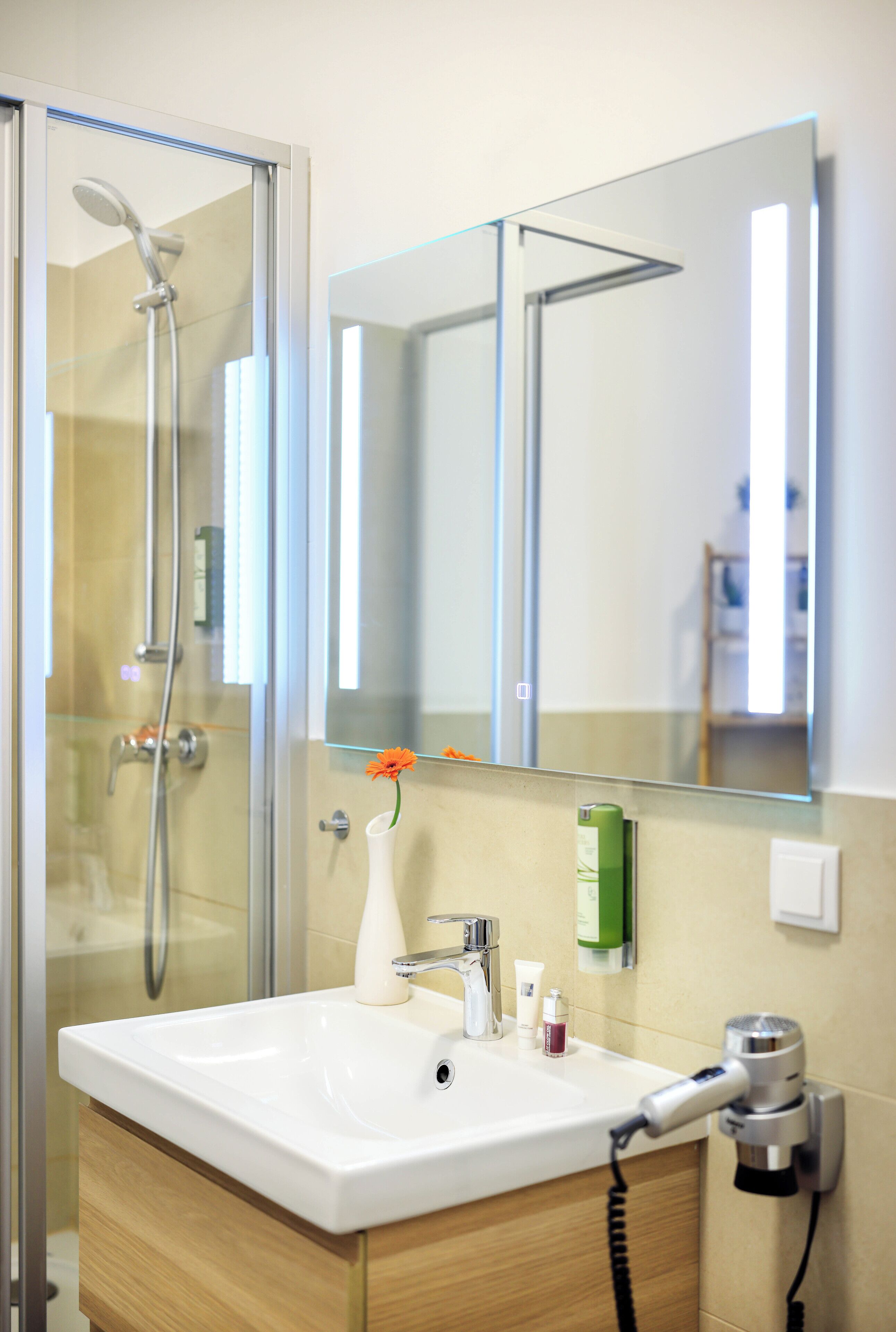 A bathroom with a glass shower enclosure, a white sink, a mirror, and a hairdryer on the wall.