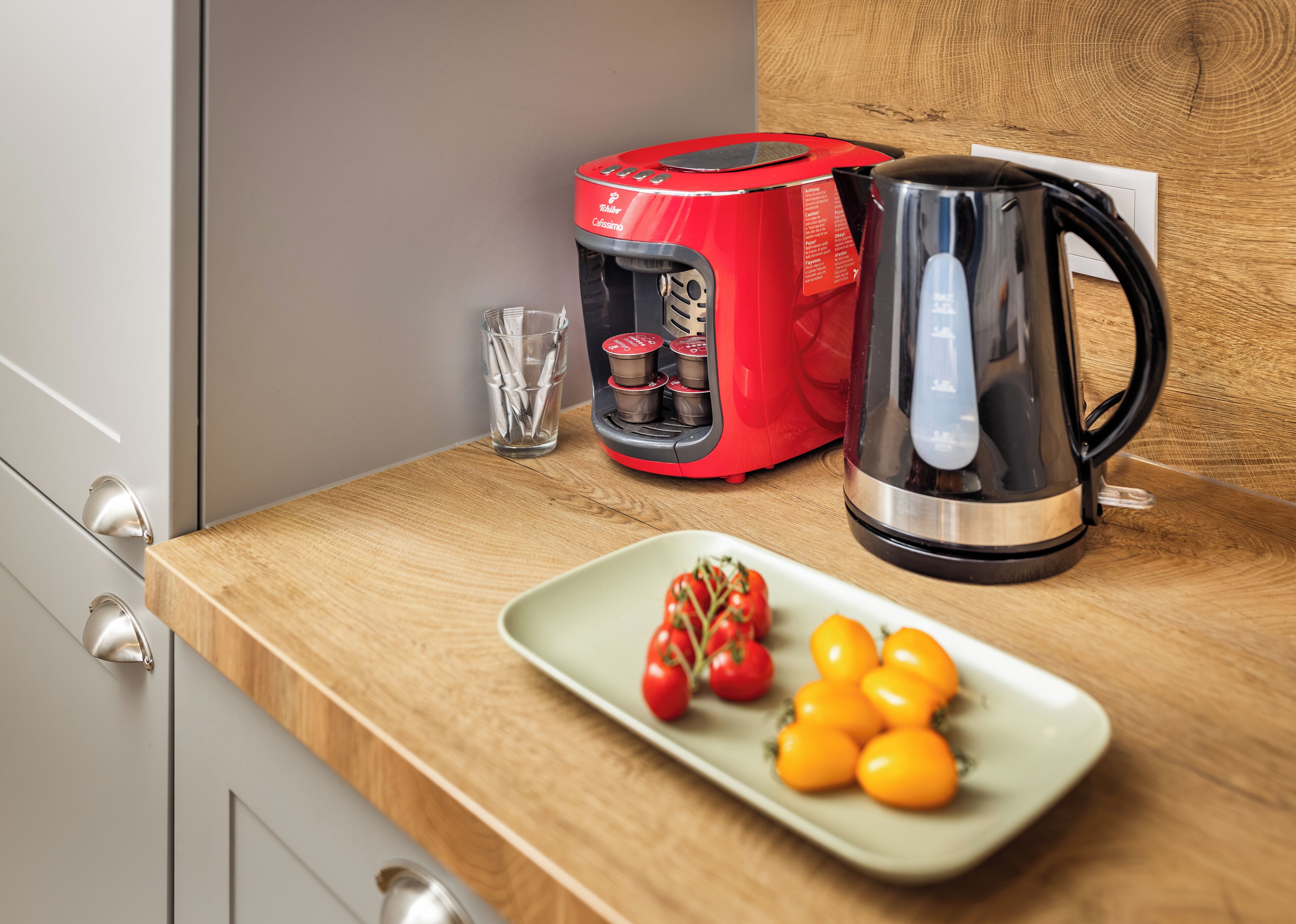 Una encimera de cocina con una cafetera roja, un vaso de agua, un hervidor eléctrico negro y un plato con tomates y tomatitos.