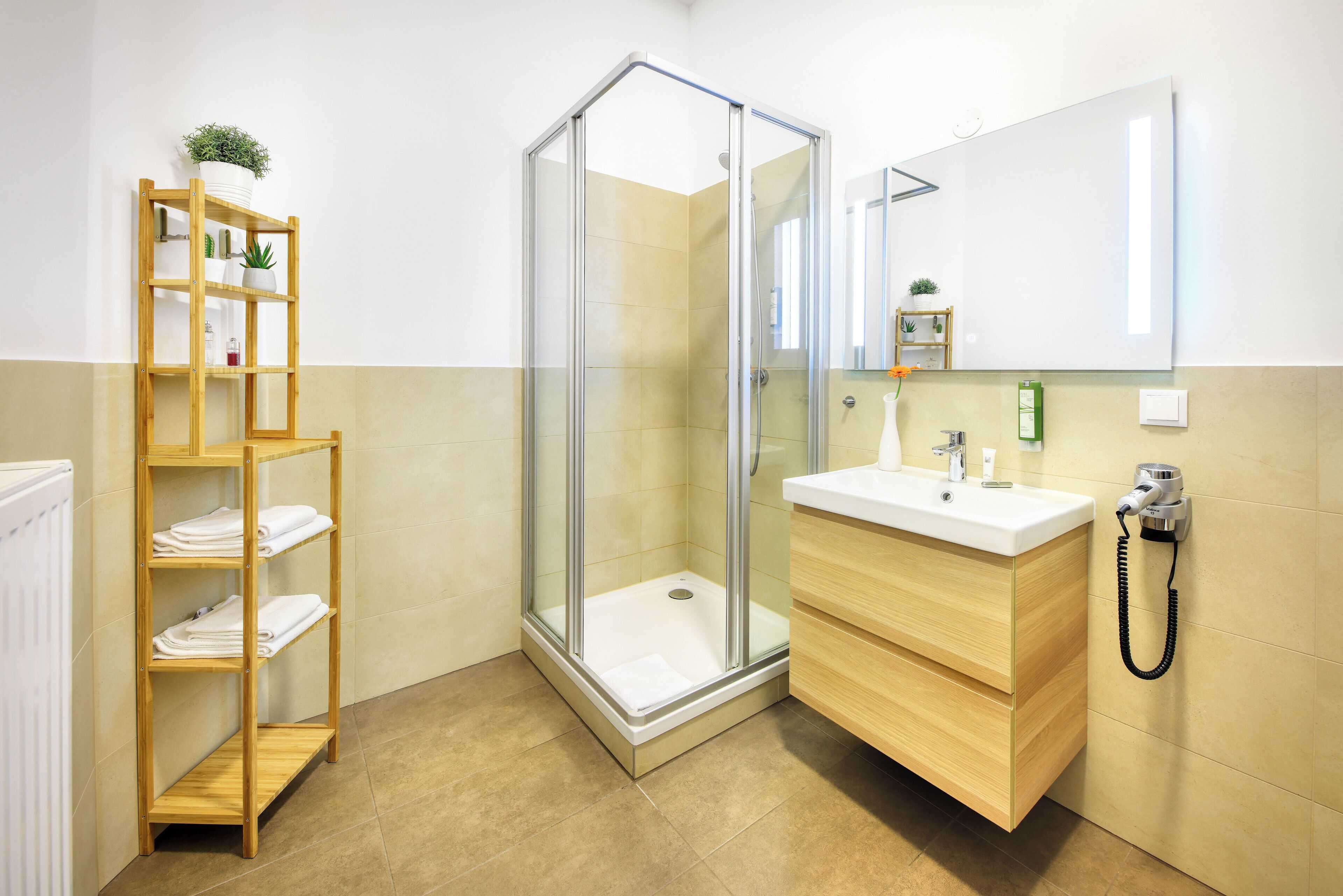 Een moderne badkamer met een glazen douchecabine, een houten plank met handdoeken, een wastafel met spiegel en een haardroger aan de muur.